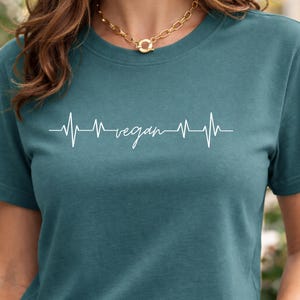 May include: Teal t-shirt featuring a white heartbeat line with the word "vegan". The top has a crew neck and short sleeves. A gold chain necklace is worn at the neckline.