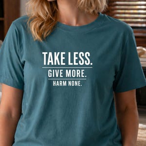 May include: Teal-colored t-shirt with the text "TAKE LESS. GIVE MORE. HARM NONE." in white block letters. The shirt has short sleeves and a crew neck. The model is wearing a gold watch.