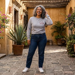 May include: A person wearing a grey long-sleeved top with the words "con amor," and dark blue jeans with white trainers. The top has a small design on the chest. The person is smiling in an outdoor setting with plants and stone paving.