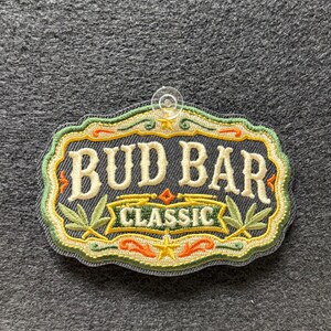 Puede incluir: Un parche bordado con fondo gris oscuro que presenta el texto "BUD BAR" en letras color crema sobre una cinta con la palabra "CLASSIC" en verde. El diseño incluye hojas verdes decorativas, detalles en naranja y una pequeña estrella amarilla.