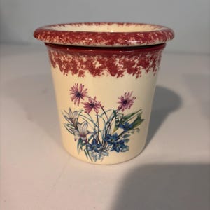 May include: A cream-colored ceramic container with a lid, featuring a floral design in purple, blue, and green. The rim and upper portion of the container are decorated with a maroon speckled pattern.