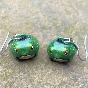 May include: A pair of green frog earrings with white spots and red eyes. The earrings are made of polymer clay and have silver hooks.