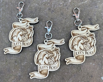 May include: Three wooden keychains with silver clasps. Each keychain features a dragon design with a banner that reads "LET'S ROLL". The dragon is holding a twenty-sided die. The keychains are made of light-colored wood.