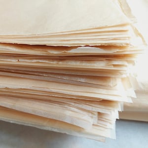 May include: A stack of thin, translucent sheets of parchment paper. The paper has a light tan color and a slightly textured surface. The edges of the sheets are uneven, suggesting they have been cut or torn. The stack is neatly arranged, with the sheets layered on top of each other.