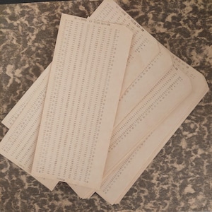 May include: A stack of off-white rectangular paper punch cards featuring columns of printed numbers and the text XI-1958 TV 57.28.107-88. The cards are arranged in an overlapping fan pattern, displaying a grid of numerical data points.