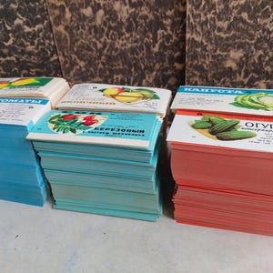 May include: Stacks of vintage seed packets featuring colorful illustrations of tomatoes, cucumbers, and other vegetables. The packets are in various colors, including blue, teal, and red, with text in a foreign language.