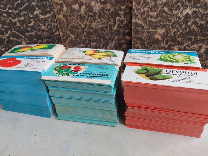 May include: Stacks of vintage seed packets featuring colorful illustrations of tomatoes, cucumbers, and other vegetables. The packets are in various colors, including blue, teal, and red, with text in a foreign language.