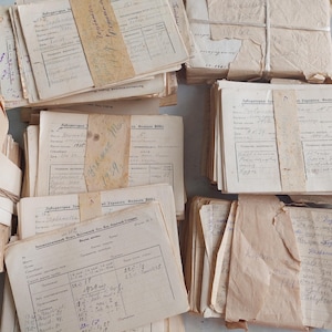 May include: A collection of vintage paper documents and laboratory forms bundled with tan paper bands and string. The off-white sheets feature printed black grids, tables, and Cyrillic text, with handwritten notes and dates in blue and black ink throughout.