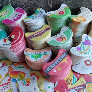 May include: A collection of crescent-shaped paper labels for beverage bottles, featuring various colorful fruit and floral illustrations, Cyrillic text, and brand names like Buratino, Moskovsky, and Vinogradny, stacked in multiple piles.