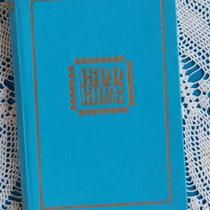 Op de afbeelding: Een fel turquoise hardcover boek met een goudkleurige rechthoekige rand en een centraal goudkleurig embleem met de tekst 1500 KHEE. De boekomslag heeft een gladde, effen afwerking met gouden letters en decoratieve accenten.