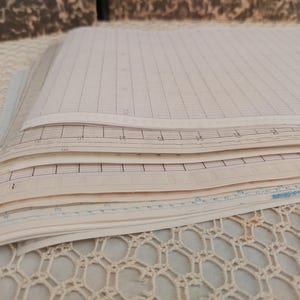 May include: A stack of off-white paper sheets featuring printed grid lines and numerical markings. The paper has a vintage appearance with visible aging and texture. Each sheet is layered, showing a sequence of technical graph paper designs.