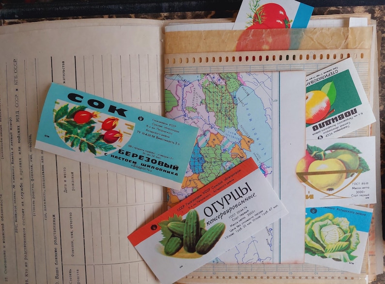 May include: An open book with vintage ephemera, including colorful labels with Cyrillic text and illustrations of fruits and vegetables. A map and other papers are also visible.