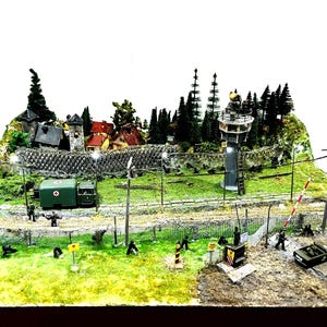 Peut inclure: Un diorama détaillé représentant une scène de poste-frontière avec une haute tour de guet grise, une clôture grillagée surmontée de barbelés, une ambulance verte, des figurines miniatures, des arbres et une barrière rayée rouge et blanche.