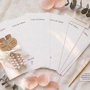 Puede incluir: Un conjunto de tarjetas de papel rectangulares blancas impresas con los nombres Laura & Fabian y la fecha 18.09.2026 con un icono de coraz&oacute;n. Dos tubos de ensayo de vidrio rellenos de peque&ntilde;os caramelos blancos y rosas, corchos y cinta blanca.