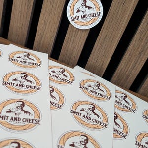 Puede incluir: Una colección de pegatinas circulares con un logotipo que muestra a una persona, un gráfico de pan circular y el texto SIMIT AND CHEESE ROTTERDAM. Las pegatinas están dispuestas en hojas blancas, con una pegatina individual colocada por separado.
