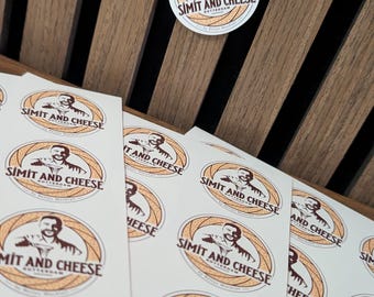 May include: A collection of circular stickers featuring a logo with a person, a circular bread graphic, and the text SIMIT AND CHEESE ROTTERDAM. The stickers are arranged on white sheets, with one individual sticker placed separately above the others.