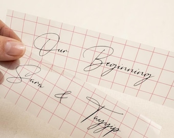 May include: Two rectangular translucent sheets featuring a thin red grid pattern. The top sheet displays the black cursive text Our Beginning, while the bottom sheet displays the black cursive text Sara & Tayyip in a matching script font.
