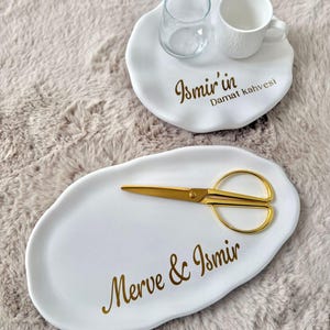 Puede incluir: Dos bandejas de cerámica blanca de forma irregular con letras doradas. La bandeja pequeña sostiene un vaso transparente y una taza blanca texturizada con Ismir'in Damat kahvesi. La bandeja ovalada grande tiene unas tijeras doradas y Merve & Ismir.
