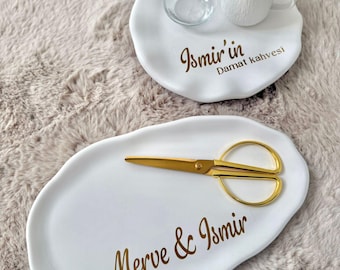 May include: Two white irregular-shaped ceramic trays with gold lettering. The smaller tray holds a clear glass and a white textured mug, inscribed with Ismir'in Damat kahvesi. The larger oval tray features gold scissors and the text Merve & Ismir.