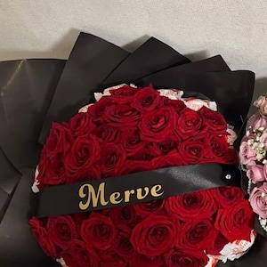 Op de afbeelding: Een boeket donkerrode rozen verpakt in zwart papier, met een zwart lint in het midden waarop de naam Merve in gouden letters is gedrukt. Witte lekkernijen zijn tussen de rozen in het boeket gestoken voor een decoratief effect.
