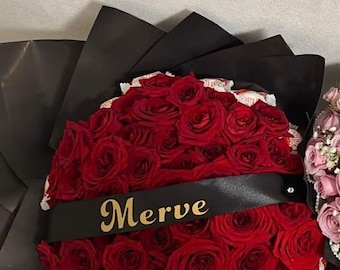 May include: A bouquet of deep red roses wrapped in black paper, featuring white Raffaello candy packaging tucked between the flowers and a black satin ribbon across the center with the name Merve printed in gold lettering.