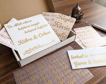 May include: A collection of custom wedding decals in a cardboard box. The decals feature gold lettering with names, dates, and phrases in various fonts. The box is open, revealing the decals on a wooden surface.