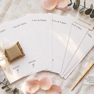 Puede incluir: Un conjunto de tarjetas de papel blanco rectangulares impresas con los nombres Laura & Fabian y la fecha 18.09.2026 con un icono de coraz&oacute;n. Una tarjeta presenta un chocolate cuadrado envuelto en dorado con una cinta blanca. Se incluye un bol&iacute;grafo dorado.