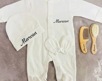 May include: A white baby clothing set featuring a onesie, hat, and mittens, all with the name "Merwan" in script. Includes a wooden comb and brush, also with the name "Merwan". The set is laid out on a soft, gray surface.