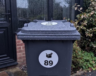 May include: A dark gray plastic wheelie bin featuring a circular white sticker on the front with a black graphic of an open cardboard box above the number 89. A smaller matching circular sticker with the number 89 is placed on the closed lid.