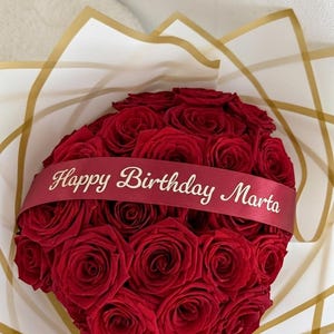 Puede incluir: Un ramo de rosas de color rojo intenso dispuestas en forma redondeada, envuelto en papel blanco con líneas geométricas doradas. Una cinta roja oscura cruza las rosas con el texto Happy Birthday Marta impreso en color dorado.
