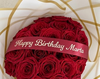 May include: A bouquet of deep red roses arranged in a rounded shape, wrapped in white paper with gold geometric lines. A dark red ribbon is draped across the center of the roses with the text Happy Birthday Marta printed in gold.