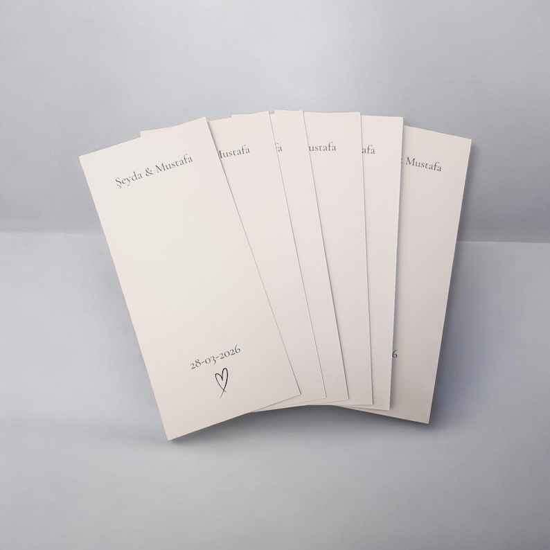 Puede incluir: Un conjunto de tarjetas de papel rectangulares de color blanco roto dispuestas en abanico. Cada tarjeta presenta el texto impreso "Şeyda & Mustafa" en la parte superior y la fecha "28-03-2026" seguida de un peque&ntilde;o coraz&oacute;n dibujado a mano.