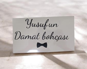 May include: A white rectangular sign with black script that reads "Yusufun Damat bohçası" and a black bow tie graphic. The sign is glossy and sits on a light-colored surface.