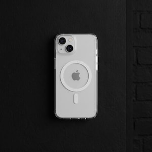 Könnte beinhalten: Eine transparente Schutzhülle für Smartphones mit einem kreisförmigen magnetischen Ausrichtungssystem auf der Rückseite. Die Hülle ist durchsichtig, zeigt das Apple-Logo des Geräts darunter und hat eine kleine vertikale Ausrichtungskerbe.