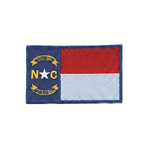 May include: A rectangular embroidered patch featuring the North Carolina state flag. It has a blue vertical stripe on the left with a white star, the letters N and C, and two yellow ribbons with the text MAY 20TH 1775 and APRIL 12TH 1776.