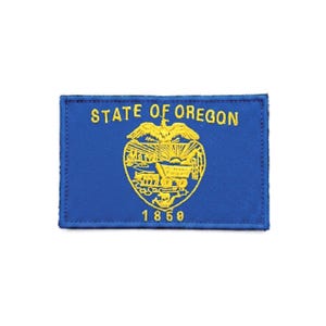 May include: A rectangular embroidered patch featuring the blue flag of the State of Oregon. The design includes yellow text reading STATE OF OREGON, a central emblem with a wagon and eagle, and the year 1859 stitched in yellow thread.