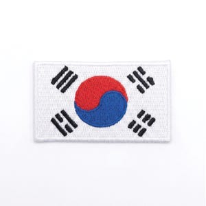 May include: Embroidered patch featuring the South Korean flag. The patch is rectangular with a white background. The flag design includes a red and blue yin-yang symbol in the center, surrounded by four black trigrams.