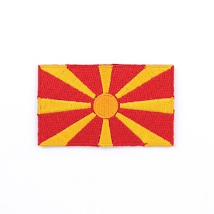 Könnte beinhalten: Gestickter Aufnäher mit der Flagge Nordmazedoniens. Der rechteckige Aufnäher hat einen roten Hintergrund mit einem gelben Sonnenstrahlendesign, das von einem zentralen Kreis ausgeht. Das Sonnenstrahlendesign besteht aus gelben Strahlen.