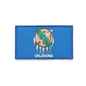May include: A rectangular embroidered patch featuring the Oklahoma state flag design. The patch has a bright blue background with a central tan shield, a crossed peace pipe and olive branch, seven white eagle feathers, and the word OKLAHOMA in white.