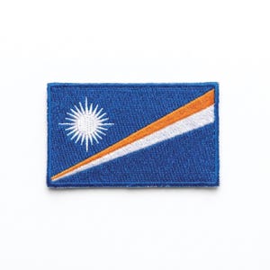 Puede incluir: Parche bordado con la bandera de las Islas Marshall. El diseño incluye un fondo azul con una estrella blanca, una franja diagonal blanca y una franja naranja. El parche es rectangular.