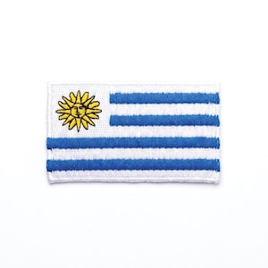 Pode incluir: Patch bordado com a bandeira do Uruguai. O design inclui um sol amarelo com um rosto no canto superior esquerdo e nove listras azuis e brancas. O patch é retangular e tem uma borda branca.