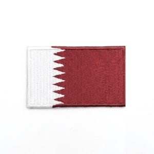 Puede incluir: Un parche rectangular con la bandera de Qatar. El parche tiene un fondo granate con un borde blanco dentado en el lado izquierdo. El borde blanco se asemeja a una serie de triángulos.