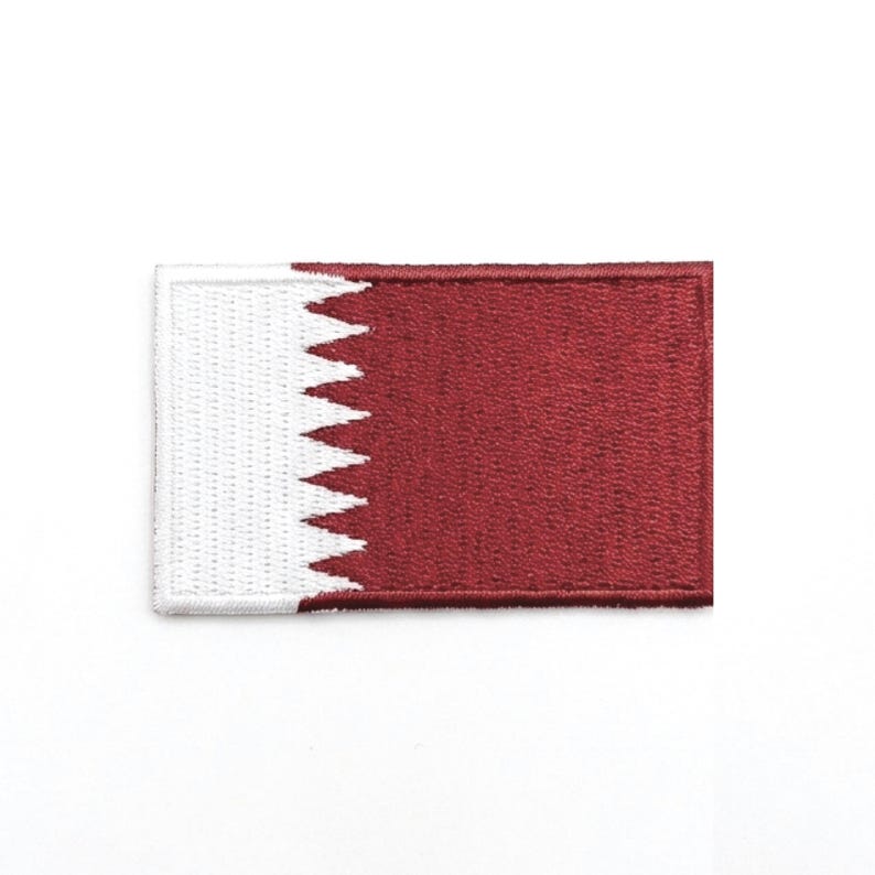 May include: A rectangular patch featuring the flag of Qatar. The patch has a maroon background with a white serrated edge on the left side. The white edge resembles a series of triangles.