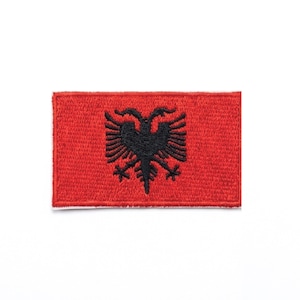 Può includere: Una toppa rettangolare rossa con un'aquila bicefala nera. La toppa ha un bordo cucito ed è probabilmente un emblema o una toppa ricamata, forse per abbigliamento o accessori.