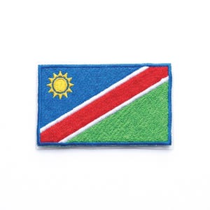 May include: Embroidered patch depicting the Namibian flag. The rectangular patch features a blue upper-left corner with a yellow sun emblem. A red stripe is bordered by white, and a green section fills the lower right corner. The patch has a white border.