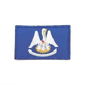 Pode incluir: Um emblema retangular bordado com fundo azul, apresentando um pelicano branco a alimentar as suas crias num ninho amarelo. Abaixo do ninho encontra-se uma faixa branca com texto. O design está centrado com uma borda costurada azul-escura.