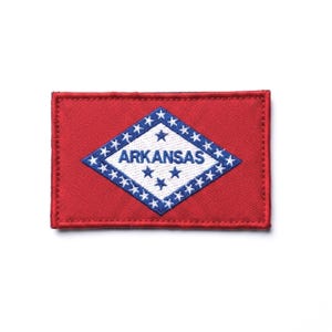 Pode incluir: Um emblema bordado retangular com o design da bandeira do estado do Arkansas. O emblema tem um fundo vermelho com um diamante branco central com borda azul, contendo o texto azul ARKANSAS e uma série de estrelas azuis.