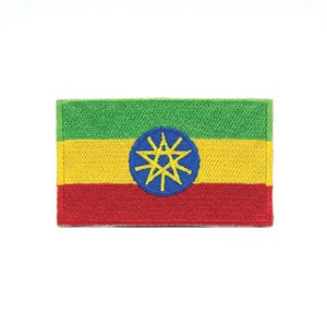 May include: Embroidered patch featuring the flag of Ethiopia. The rectangular patch displays horizontal stripes of green, yellow, and red. A blue circle in the center contains a yellow starburst design. The patch is likely for clothing or accessories.