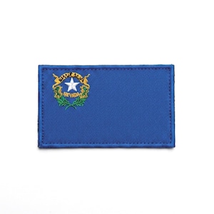 Peut inclure: Un écusson rectangulaire brodé représentant le drapeau bleu du Nevada. Dans le coin supérieur gauche, une étoile blanche est centrée dans une bannière dorée et des branches de sauge vertes, avec le mot NEVADA brodé en or sous l'étoile.