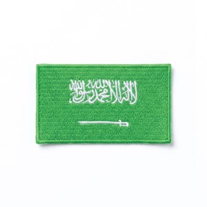 Puede incluir: Un parche rectangular verde con la bandera de Arabia Saudita. El diseño de la bandera incluye una inscripción blanca y una espada blanca. El parche probablemente está hecho de tela y está diseñado para ser cosido o planchado en ropa u otros artículos.